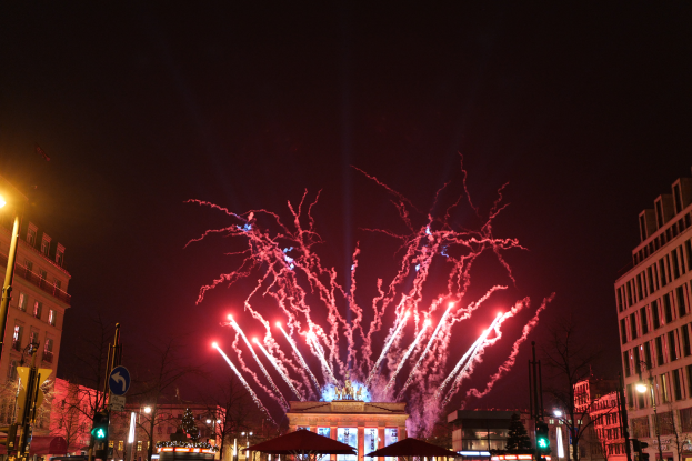 Eine belebte Stadtstraße in Berlin an Silvester, voller Menschen, Fahrzeuge und Gebäude, beleuchtet von Feuerwerk und Lichtern, die eine festliche Atmosphäre schaffen.