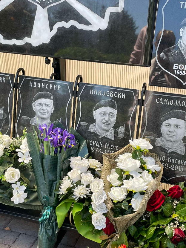 Ein Strauß aus farbenfrohen Blumen, darunter weiß, rot und lila, auf einem Gehweg neben einem Schild, mit mehreren Gedenktafeln im Hintergrund an einer Wand befestigt.