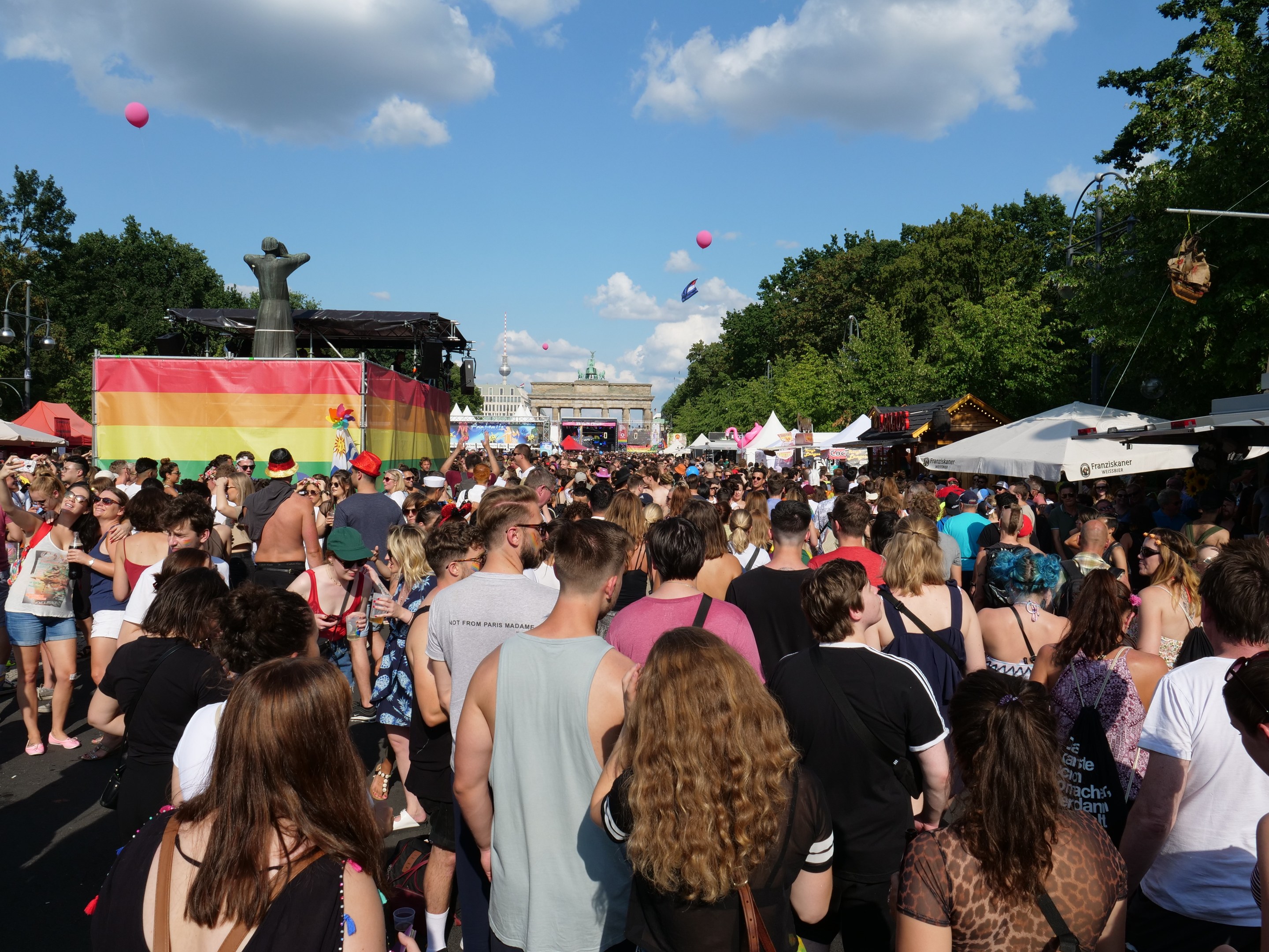 Eine große Menschenmenge geht eine Straße mit Zelten, Bäumen, Pfählen, Lichtern und einer Statue entlang während des Christopher Street Day Festivals in Berlin, mit Gebäuden, Wolken und Ballons im Hintergrund.