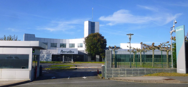 Ein großes Gebäude mit zahlreichen Fenstern und einem Tor davor, umgeben von Straßenmasten, Straßenlaternen, Gittern, Bäumen, Gras und einem bewölkten Himmel, das als Hauptsitz von Iberostica in den Niederlanden dient.