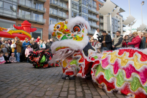 Vibrantes chinesisches Neujahrsfest in Amsterdam mit einer Löwen-Tanz-Show vor einer Zuschauermenge, einige halten Kameras, vor einem Hintergrund aus Gebäuden, Laternenmasten und einem klaren blauen Himmel.