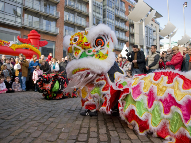 Vibrantes chinesisches Neujahrsfest in Amsterdam mit einer Löwen-Tanz-Show vor einer Zuschauermenge, einige halten Kameras, vor einem Hintergrund aus Gebäuden, Laternenmasten und einem klaren blauen Himmel.