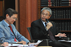 Drei Personen sitzen auf Stühlen, eine hält ein Mikrofon, die anderen halten Stifte und Papiere, im Hintergrund ein Bücherregal.