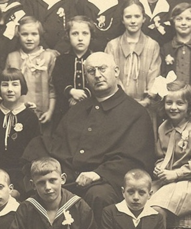 Ein Schwarz-Weiß-Foto eines ernst aussehenden Mannes, der in der Mitte sitzt und von einer Gruppe von Kindern umgeben ist, die alle bewundernd zu ihm aufschauen.