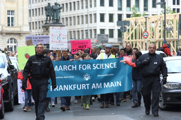 Eine Gruppe von Menschen marschiert auf einer Straße und hält ein "March for Science Frankfurt am Main"-Schild hoch, mit Autos, Gebäuden, Statuen, Laternenmasten, Schildern und Bäumen im Hintergrund.