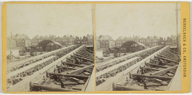 Schwarz-weiß-Foto einer Zugschiene mit Gebäuden im Hintergrund, Text 'Merkelbach & Co, Co. Amsterdam' rechts und verstreuten Holzobjekten im Vordergrund.