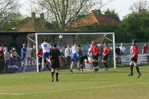 Fußballspieler spielen auf einem Feld mit einem Tor und Zuschauern dahinter, mit Bäumen und Häusern im Hintergrund.