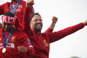 Liverpool-Trainer Jürgen Klopp feiert mit seinen Spielern den Sieg in der UEFA Champions League, umgeben von zwei medaillentragenden Individuen, mit dem Himmel im Hintergrund.