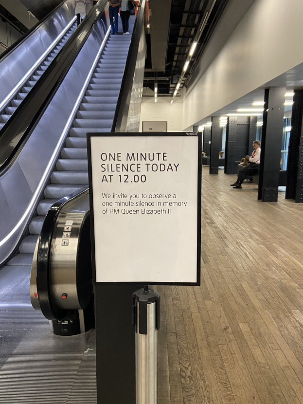 Eine Rolltreppe im Flughafen mit einem Schild, auf dem "Eine Minute Stille heute" steht, sowie ein paar Menschen darauf und beleuchtete Decke im Hintergrund.