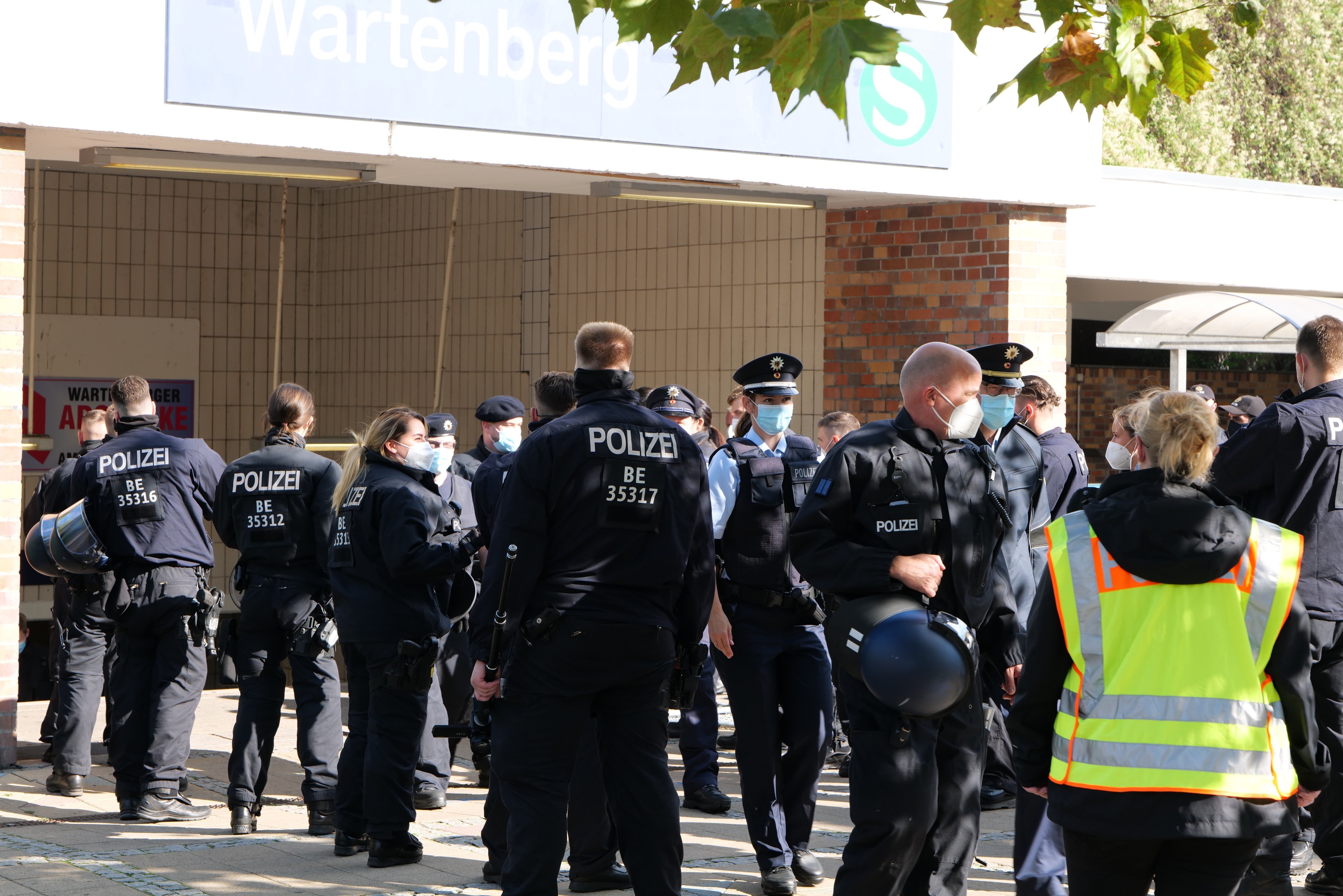 Eine Gruppe von Polizisten in Uniform steht vor einem Gebäude mit einem Schild und Bäumen dahinter, einige tragen Mützen und Masken, andere halten Helme, mit einer Texttafel an der Wand des Gebäudes.