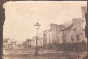 Alte Schwarz-Weiß-Fotografie einer Straßenlaterne an einer Kreuzung, flankiert von Gebäuden unter einem klaren Himmel.