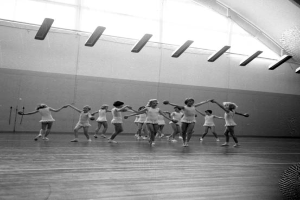 Eine Gruppe junger Mädchen in einem Tanzstudio, die sich an den Händen halten und auf dem Boden tanzen, mit einem Ball auf der rechten Seite, Lampen an der Decke und einem Objekt an der Wand im Hintergrund, alles in Schwarz-Weiß dargestellt.