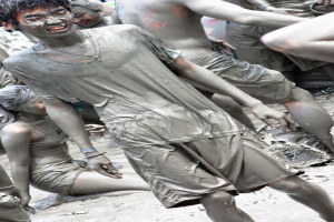 Ein Junge lächelt und geht im Schlamm, mit Schlamm auf seiner Kleidung und seinem Körper, während im Hintergrund andere Personen mit Schlamm an sich zu sehen sind.