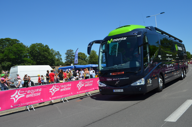 Ein schwarzer und grüner Bus fährt eine Straße entlang neben einer Menschenmenge, einige tragen Mützen, mit einem Banner auf der linken Seite und Bäumen unter einem klaren blauen Himmel im Hintergrund.