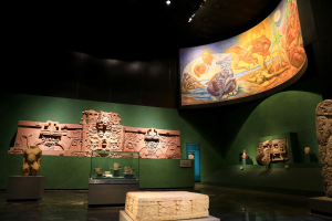 Ein Museumsraum mit verschiedenen Artefakten, darunter Skulpturen, Gegenstände auf dem Tisch, eine Texttafel, eine Wandmalerei und Deckenleuchten im Anthropologischen Museum von Mexiko-Stadt.