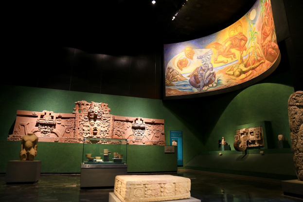 Ein Museumsraum mit verschiedenen Artefakten, darunter Skulpturen, Gegenstände auf dem Tisch, eine Texttafel, eine Wandmalerei und Deckenleuchten im Anthropologischen Museum von Mexiko-Stadt.