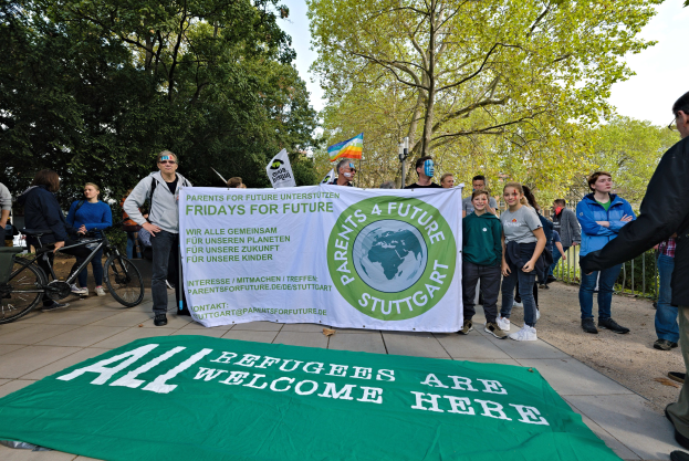 Eine Gruppe von Menschen mit einer Tafel, auf der "All Refugees Are Welcome Here" steht und einer Flagge, sowie Fahrrädern, einem Zaun, einem Straßenschild, einem Schild, Bäumen und einem bewölkten Himmel im Hintergrund.