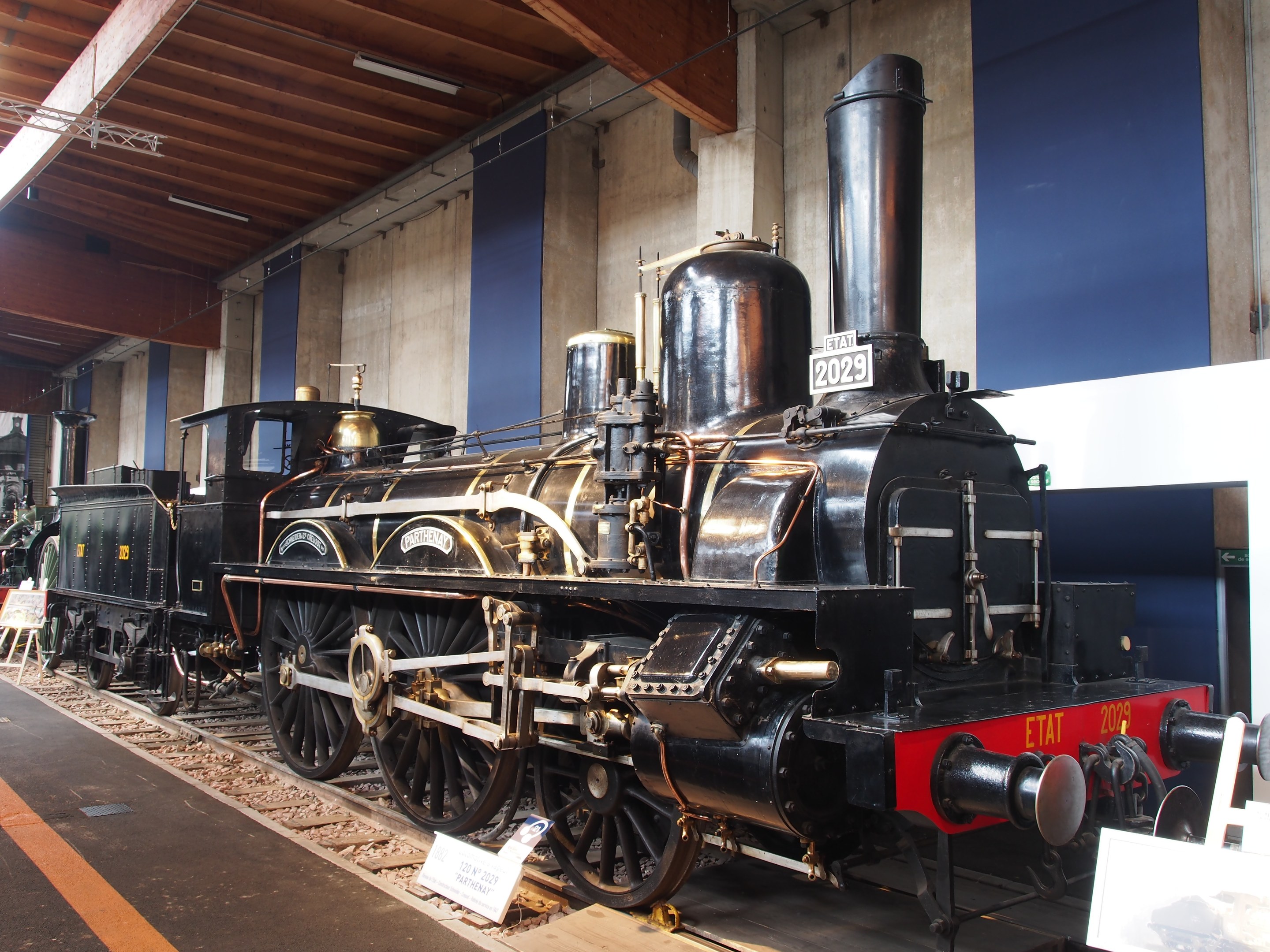 Alte Dampflokomotive im Museum ausgestellt, umgeben von Eisenbahnschienen und Informationsschildern, mit beleuchteter Decke im Hintergrund.