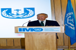 Eine Person steht hinter einem Podium mit einem Mikrofon, Brille und Text, spricht, mit einer Wandtafel mit Logo und einer Flagge im Hintergrund und einer Wand und einem Boden, die die Szene vervollständigen.