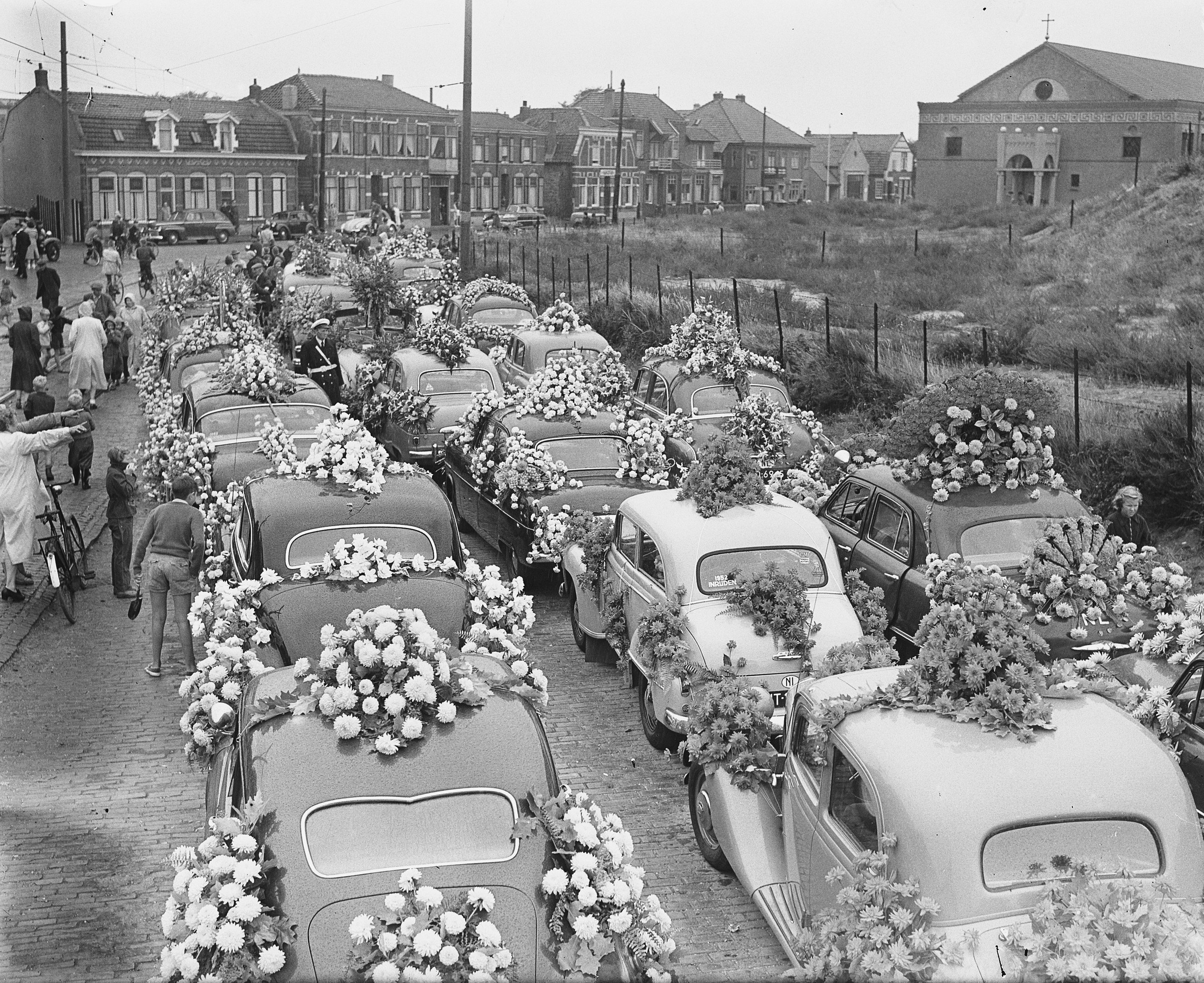 Ein Schwarz-Weiß-Bild einer Beerdigungsprozession mit Autos, die mit Blumenbouquets geschmückt sind, Menschen mit Fahrrädern und Gebäuden im Hintergrund.
