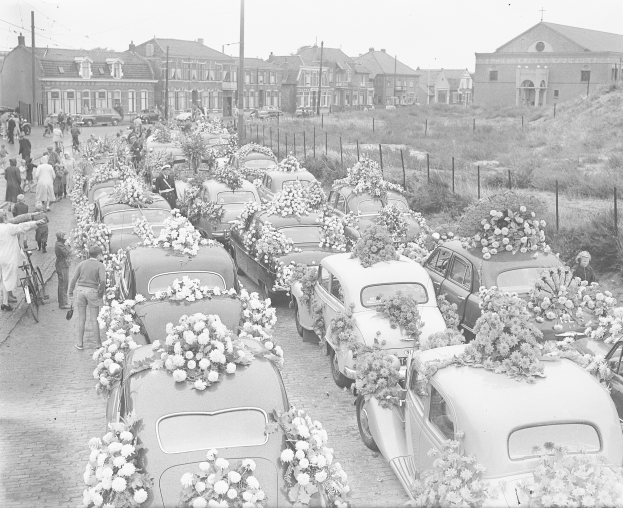 Ein Schwarz-Weiß-Bild einer Beerdigungsprozession mit Autos, die mit Blumenbouquets geschmückt sind, Menschen mit Fahrrädern und Gebäuden im Hintergrund.