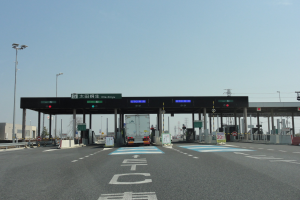 Eine Mautstation mit einem davor geparkten Lastwagen, neben einer Straße mit weißen und blauen Linien, Laternenmasten und umliegenden Gebäuden, Bäumen und einem klaren Himmel.