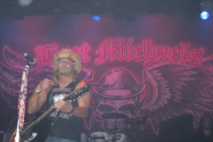 Eine Musikkonzertszene mit Scheinwerfern oben, einem Banner im Hintergrund, Drogen in der Mitte unten, einer Person in der rechten unteren Ecke und einem Gitarristen mit Mikrofon links.