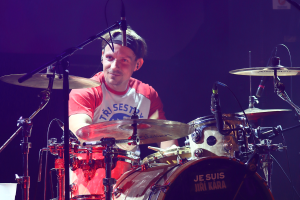 Ein Mann spielt Schlagzeug auf der B├╝hne bei einem Konzert, trägt ein rotes und weißes T-Shirt und lacht, mit mehreren Mikrofonen vor ihm.