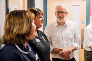 Eine Gruppe von Menschen, die in einem Raum mit einer Tür im Hintergrund und einem Anschlagbrett mit Papieren an der linken Seite stehen und sprechen.