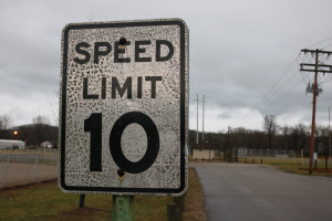 Geschwindigkeitsbegrenzungstafel am Straßenrand mit umliegender Wiese, Strommästen, einem Zaun, Häusern, Bäumen und einem bewölktem Himmel.