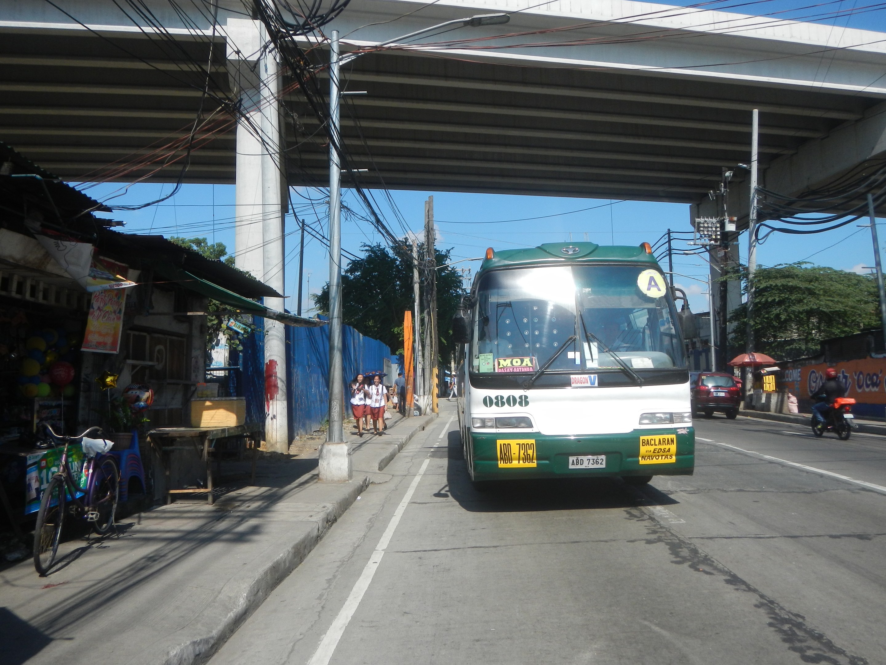 Ein grüner und weißer Bus fährt auf einer Straße mit Strommasten und -leitungen entlang, vorbei an Geschäften und Ständen, mit Menschen auf dem Gehweg und Fahrrädern in der Nähe, während Bäume und eine Brücke im Hintergrund unter einem klaren blauen Himmel zu sehen sind.