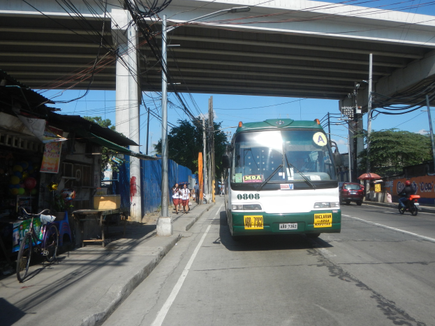 Ein grüner und weißer Bus fährt auf einer Straße mit Strommasten und -leitungen entlang, vorbei an Geschäften und Ständen, mit Menschen auf dem Gehweg und Fahrrädern in der Nähe, während Bäume und eine Brücke im Hintergrund unter einem klaren blauen Himmel zu sehen sind.