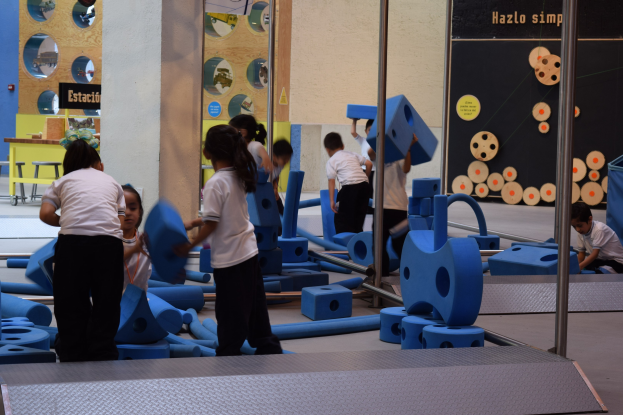 Kinder beim Spielen in einer Turnhalle mit einigen, die Gegenstände halten, eine Texttafel an der Wand und Stangen auf der rechten Seite.
