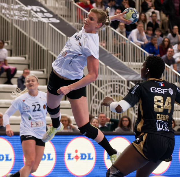Eine Gruppe von Frauen, die auf einem Handballfeld spielt, mit einem Ball in der Luft, Zuschauern auf Treppen und Geländern im Hintergrund und einem Schild mit der Aufschrift "Handball-Weltmeisterschaft 2019 - Tag 2".