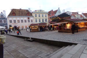 Ein belebter Weihnachtsmarkt in Tallinn, Estland mit Menschen um geschmückte Stände, festliche Lichter, bewölkter Himmel, Treppe nach oben und Topfpflanzen im Bereich.