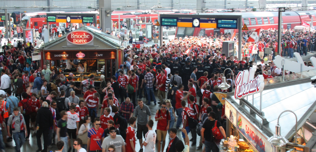 Eine große Menschenmenge, die durch einen belebten Bahnhof geht, mit einem Zug auf den Schienen im Hintergrund, umgeben von Geschäften, Texttafeln, Pfählen, Lampen und anderen Bahnhofsobjekten.