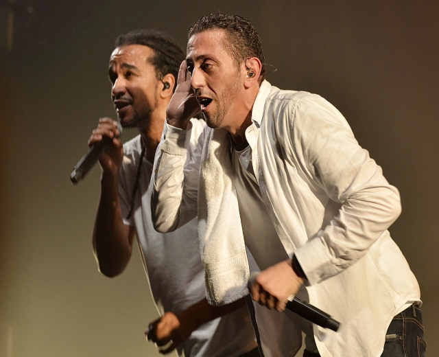 Zwei Männer in legerer Kleidung performen auf der Bühne Seite an Seite, jeder hält ein Mikrofon in der Hand.