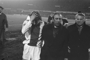 Eine Gruppe von Männern in Not steht zusammen auf einem Fußballfeld, mit einem Stadion im Hintergrund, dargestellt in Schwarz-Weiß.