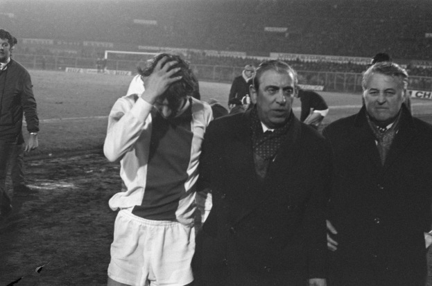 Eine Gruppe von Männern in Not steht zusammen auf einem Fußballfeld, mit einem Stadion im Hintergrund, dargestellt in Schwarz-Weiß.