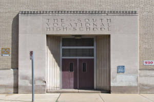 Das Südliche Berufsbildungszentrum mit einer Tür, Schilder an der Wand und einem Weg, der zur Eingangstür führt.