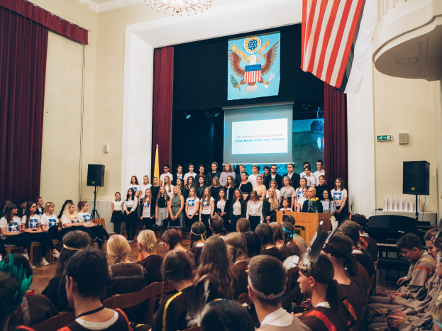 Eine Gruppe von Menschen steht auf einem Podium mit einem Mikrofon, flankiert von Fahnen und Lautsprechern, und spricht eine sitzende Menge an, während im Hintergrund ein Bildschirm, Vorhänge und ein Kronleuchter zu sehen sind.