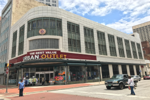 Eine belebte Stadtstraße mit Fahrzeugen, Fußgängern, hohen Gebäuden und einem Schild mit der Aufschrift "Best Value Urban Outlet" unter einem bewölkten Himmel.