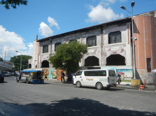 Eine belebte Stadtstraße mit Fahrzeugen, Fußgängern, Bäumen, Laternen, einem großen Gebäude mit einer Wandmalerei, einer Brücke im Hintergrund und einem bewölkten Himmel.
