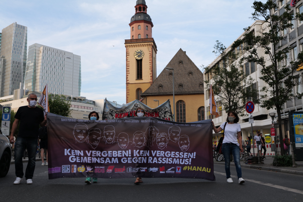 Gruppe von maskierten Menschen auf der Straße mit einem Banner, daneben ein geparktes Auto, im Hintergrund Gebäude und ein Kirchturm unter einem klaren blauen Himmel.
