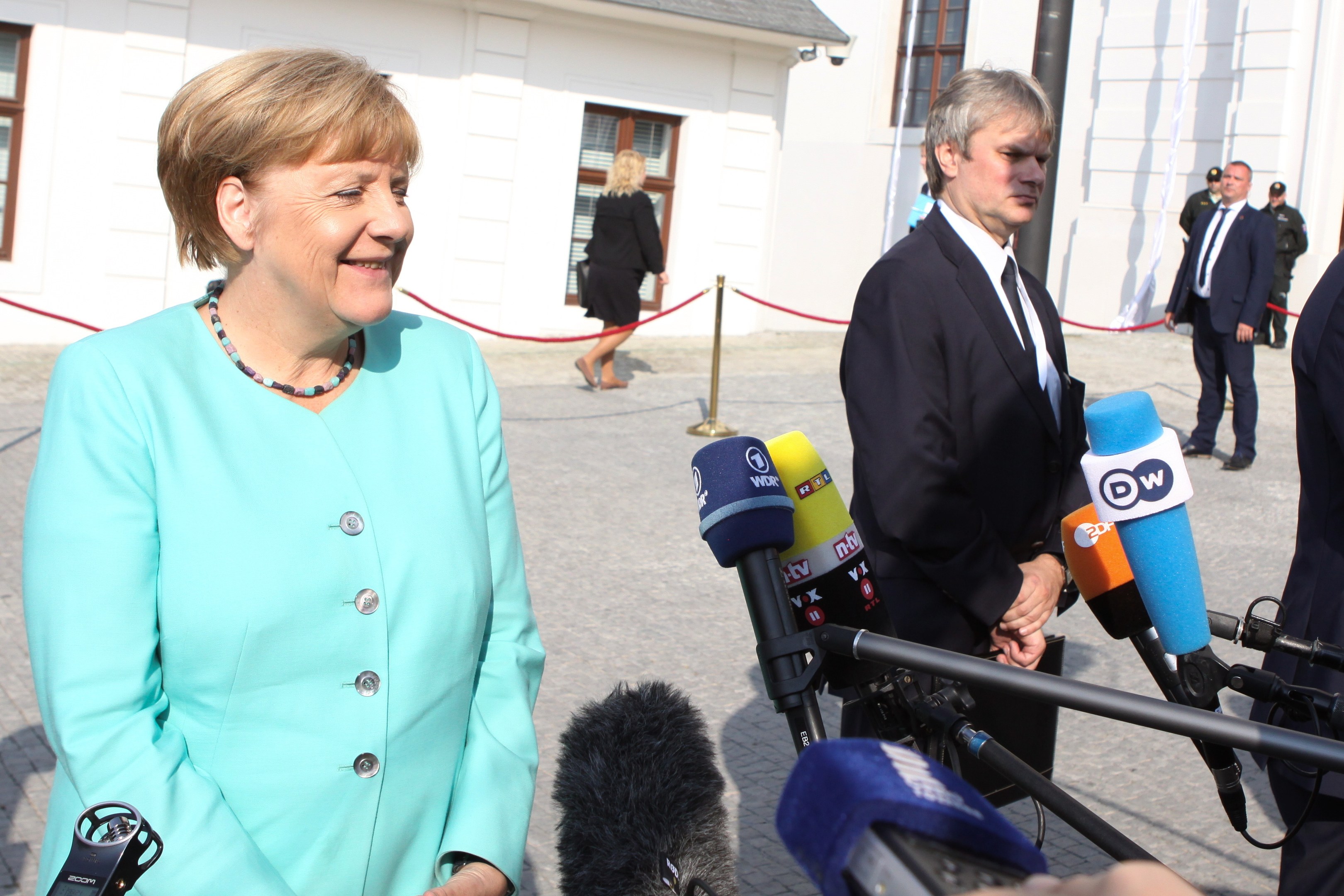 Bundeskanzlerin Angela Merkel spricht zu Reportern vor einem Gebäude mit Fenstern, umgeben von Menschen mit Mikrofonen und einer Seilabsperrung bei bewölktem Himmel.
