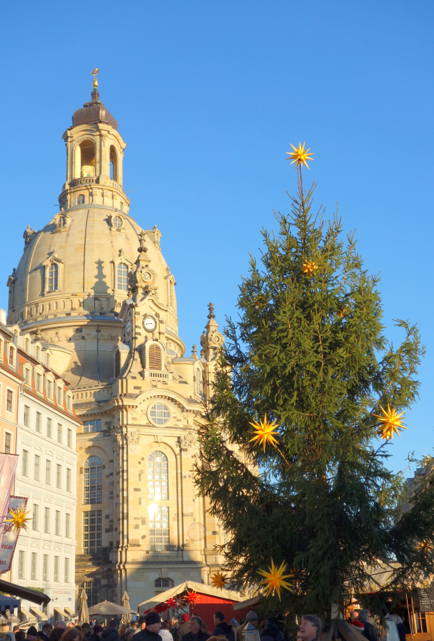 Ein geschäftiger Weihnachtsmarkt in Dresden, Deutschland, mit einem großen Weihnachtsbaum vor einer Kirche, vielen Menschen drumherum und einer Tafel mit Text auf der linken Seite, unter einem sichtbaren Himmel.