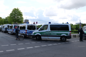 Eine Gruppe von Polizeifahrzeugen, die entlang einer Straße geparkt sind, mit einem Radfahrer im Vordergrund und Gebäuden, Bäumen, Schildern im Hintergrund.