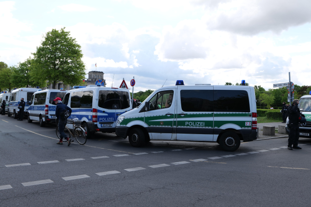 Eine Gruppe von Polizeifahrzeugen, die entlang einer Straße geparkt sind, mit einem Radfahrer im Vordergrund und Gebäuden, Bäumen, Schildern im Hintergrund.