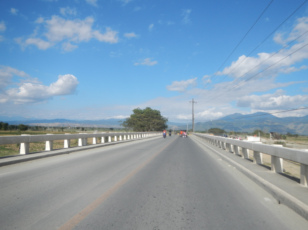 Eine Autobahn mit Motorradfahrern, umgeben von Geländern, Strommästen, Bäumen, Hügeln und einem bewölkten Himmel.