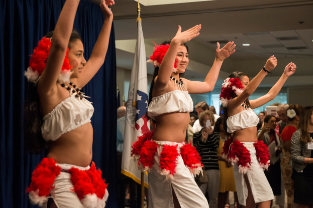 Eine Gruppe junger Frauen in weißen und roten Outfits tanzt vor einer Menge, mit einer Fahne, Vorhängen und Deckenlampen im Hintergrund.